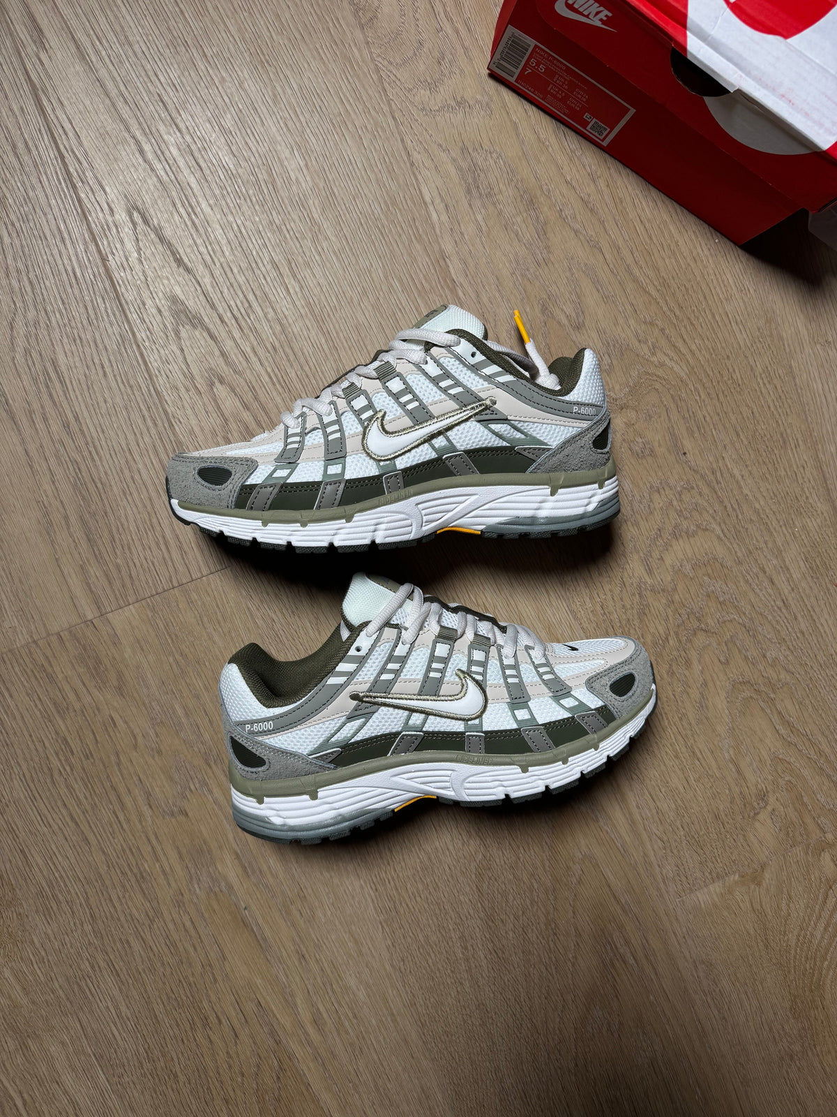 A pair of Nike P-6000 sneakers in white and gray, placed on a light brown wooden floor next to a red Nike shoebox.